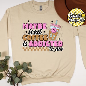 Puede incluir: Una sudadera beige con el texto "MAYBE iced COFFEE is ADDICTED to me" en letras rosas y marrones. Un vaso de caf&eacute; helado de dibujos animados con una cara sonriente est&aacute; por encima del texto.