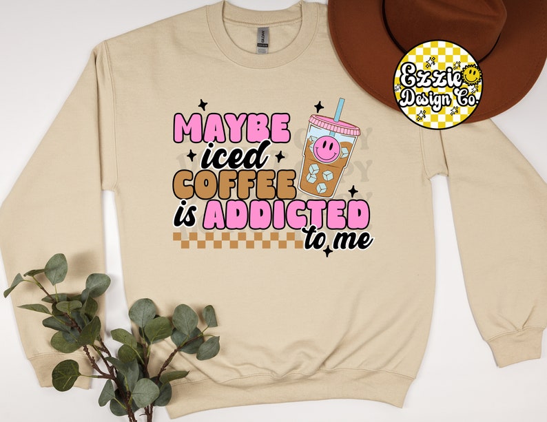 Puede incluir: Una sudadera beige con el texto "MAYBE iced COFFEE is ADDICTED to me" en letras rosas y marrones. Un vaso de caf&eacute; helado de dibujos animados con una cara sonriente est&aacute; por encima del texto.