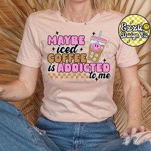 Puede incluir: Una camiseta rosa con el texto "Maybe iced coffee is addicted to me" en letras rosas y marrones. Un vaso de caf&eacute; helado de dibujos animados con una cara sonriente est&aacute; al lado del texto.