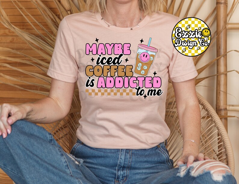Puede incluir: Una camiseta rosa con el texto "Maybe iced coffee is addicted to me" en letras rosas y marrones. Un vaso de caf&eacute; helado de dibujos animados con una cara sonriente est&aacute; al lado del texto.