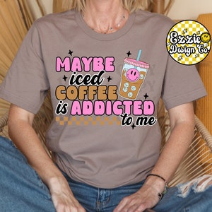 Puede incluir: Una camiseta gris con el texto "Maybe iced coffee is addicted to me" en letras rosas y marrones. Un vaso de caf&eacute; helado de dibujos animados con una cara sonriente est&aacute; al lado del texto.