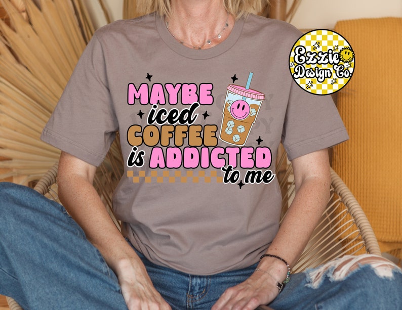 Puede incluir: Una camiseta gris con el texto "Maybe iced coffee is addicted to me" en letras rosas y marrones. Un vaso de caf&eacute; helado de dibujos animados con una cara sonriente est&aacute; al lado del texto.