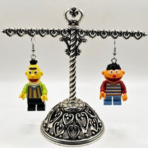 以下が含まれることがあります： 銀製のイヤリングスタンドに、イヤリングが2組飾られています。1組は緑色のシャツを着た黄色いセサミストリートのキャラクター、もう1組は青色のシャツを着た赤いセサミストリートのキャラクターです。