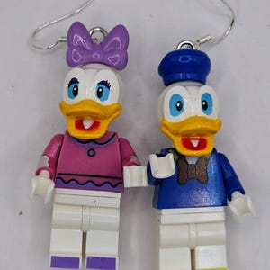 May include: A pair of dangle earrings featuring Lego minifigures of Donald and Daisy Duck. Donald is wearing a blue sailor shirt and hat, while Daisy is wearing a pink shirt with a purple bow.
