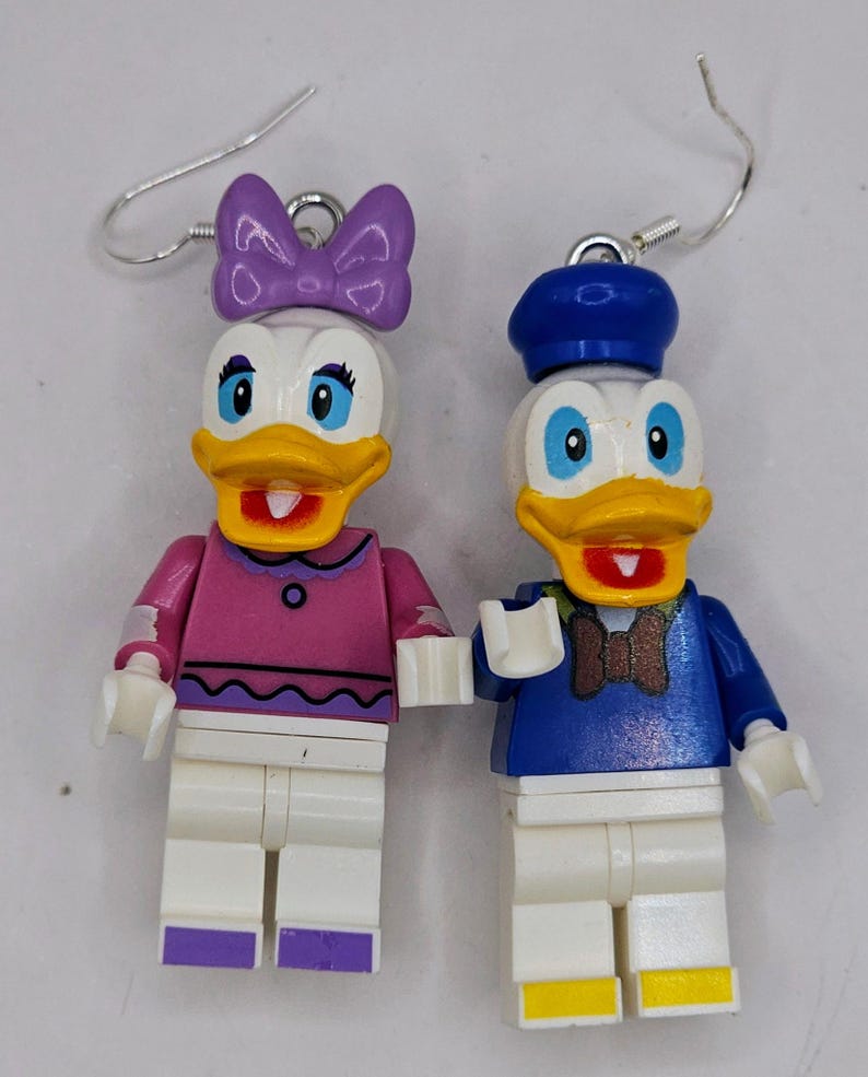 May include: A pair of dangle earrings featuring Lego minifigures of Donald and Daisy Duck. Donald is wearing a blue sailor shirt and hat, while Daisy is wearing a pink shirt with a purple bow.