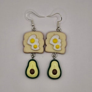 May include: A pair of dangle earrings featuring a miniature avocado and a slice of toast with fried eggs. The avocado is green with a brown pit and the toast is beige with yellow eggs.