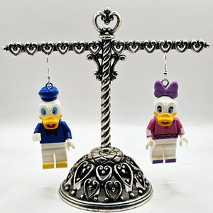 May include: A pair of dangle earrings featuring a white and blue Donald Duck Lego minifigure and a white and pink Daisy Duck Lego minifigure. The earrings are hanging from a silver heart-shaped earring stand.