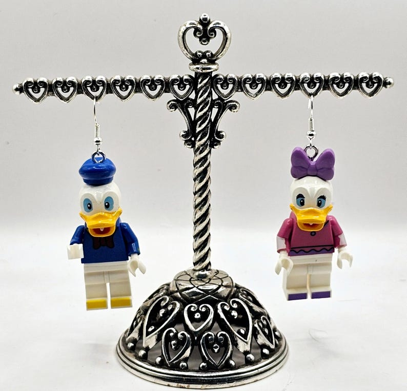 May include: A pair of dangle earrings featuring a white and blue Donald Duck Lego minifigure and a white and pink Daisy Duck Lego minifigure. The earrings are hanging from a silver heart-shaped earring stand.