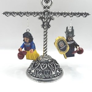 May include: Silver-toned earring display stand with two character earrings. One earring features a figure in a blue top and yellow skirt, holding a red apple. The other earring has a figure in a black outfit and a gold shield.