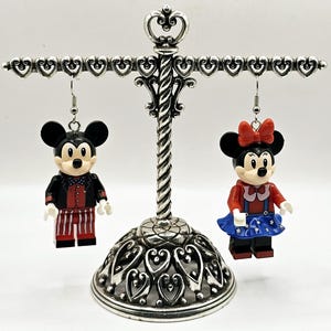 May include: A pair of dangle earrings featuring Lego minifigures of Mickey and Minnie Mouse dressed in red, white, and blue patriotic outfits. The earrings are hanging from a silver heart-shaped earring stand.