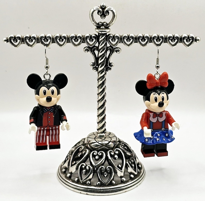 May include: A pair of dangle earrings featuring Lego minifigures of Mickey and Minnie Mouse dressed in red, white, and blue patriotic outfits. The earrings are hanging from a silver heart-shaped earring stand.