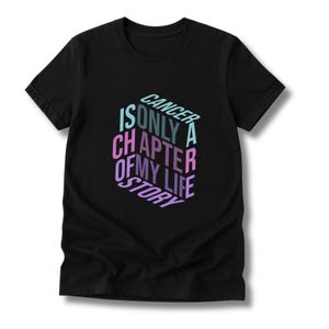 May include: Black t-shirt with the text "CANCER IS ONLY A CHAPTER OF MY LIFE STORY" in a colorful, 3D design. The text is in shades of blue, pink, and purple. The shirt is a short-sleeved crew neck.