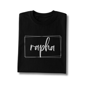 May include: Black crew neck t-shirt with the word "rapha" in white script inside a white rectangular frame. The t-shirt is folded neatly, showcasing the design prominently.