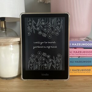May include: A Kindle e-reader with a clear protective case displays a floral design and the text "I will go to secret gardens in my mind." The e-reader is on a wooden surface, next to a stack of books and a pink flower pot.