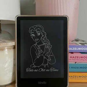 May include: A Kindle e-reader in a clear case shows a white outline of a woman with a book, and the words "Tale as Old as Time." It sits on wood, with books and a pink plant pot behind it. The e-reader is a popular device for reading.