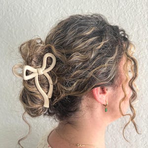 May include: A woman with curly brown hair is wearing a cream-colored bow hair clip. The hair clip is in the shape of a bow and is made of a smooth, plastic-like material. The woman is also wearing a gold necklace and a pair of gold earrings with green stones.