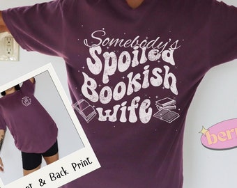 Può includere: Una maglietta viola con il testo "Somebody's Spoiled Bookish Wife" in bianco con un font in stile vintage. La maglietta ha una piccola grafica di un libro sul davanti e una grafica più grande di una pila di libri sul retro.