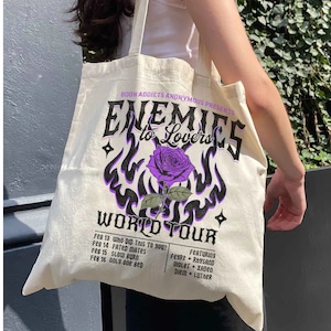 May include: A white canvas tote bag with a purple and black graphic design featuring a rose and the text "Enemies to Lovers World Tour".