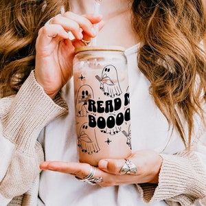 Peut inclure: Un gobelet en verre transparent avec un couvercle en bambou et une paille. Le gobelet présente un motif avec des fantômes de dessins animés et les mots "Read & Boo". La boisson à l'intérieur est brun clair.