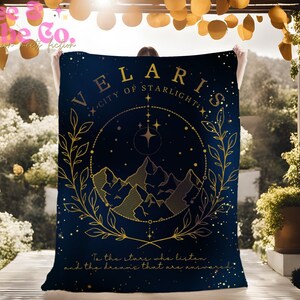 K&ouml;nnte beinhalten: Eine marineblaue Kuscheldecke mit einem goldenen Design, das eine Bergkette, Sterne und den Text "VELARIS - CITY OF STARLIGHT" zeigt. Der Text "To the stars who listen and the dreams that are whispered" befindet sich am unteren Rand der Decke.