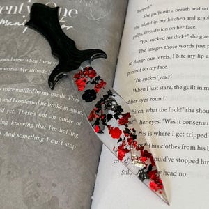 May include: A black and red dagger-shaped bookmark with glitter and floral accents, resting on an open book. The bookmark has a black handle and a clear resin blade filled with red, black, and silver glitter and floral elements.