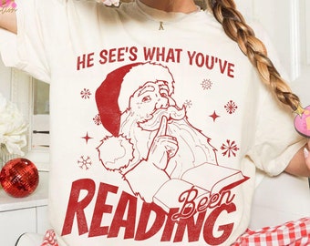 Könnte beinhalten: Cremefarbenes T-Shirt mit roter Grafik von Weihnachtsmann beim Lesen eines Buches, mit dem Text "HE SEE'S WHAT YOU'VE BEEN READING". Dazu eine rot-weiß karierte Pyjamahose. Festliche Weihnachtskleidung.
