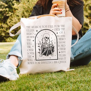 Peut inclure: Un sac fourre-tout blanc avec du texte noir et un graphique d'un squelette lisant un livre. Le texte dit "La raison pour laquelle vous tombez amoureux du méchant plutôt que du héros d'une histoire est que le héros choisira le monde plutôt que son amant, mais le méchant le brûlera pour avoir insulté sa reine."