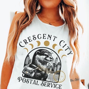 May include: White vest top with a graphic of an otter carrying a postal bag, and the text "CRESCENT CITY POSTAL SERVICE". The design includes a cityscape and crescent moon phases.
