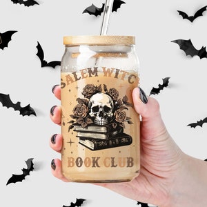 Peut inclure: Un gobelet en verre avec un couvercle en bambou et une paille en acier inoxydable. Le gobelet présente un motif avec un crâne, des roses et le texte "Salem Witch Book Club". Des décorations de chauves-souris noires sont éparpillées.