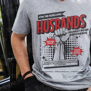 以下が含まれることがあります： コミック風のグラフィックがプリントされたグレーのTシャツ。"Husbands Join the Rebellion Against Book Boyfriends Est Out of Necessity Support Group Meets Every Sunday Because we will never be 6's, broody, immortal, or emotionally damaged in the right way."と書いてあります。