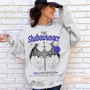 Może przedstawiać: Szara bluza z napisem "The Shadowsinger" w niebieskiej gotyckiej czcionce. Czarno-szara grafika miecza, skrzydeł i kryształów jest wyśrodkowana pod tekstem. Na bluzie wydrukowano również słowa "Illyrian Warrior" i "Spy Master of the Night Court".