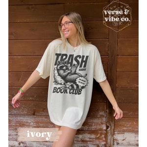Puede incluir: Una mujer lleva una camiseta de color crema con una ilustraci&oacute;n en blanco y negro de un mapache leyendo un libro. La camiseta dice "Trash Reader Book Club" y tiene un bocadillo que dice "The dirtier the better".