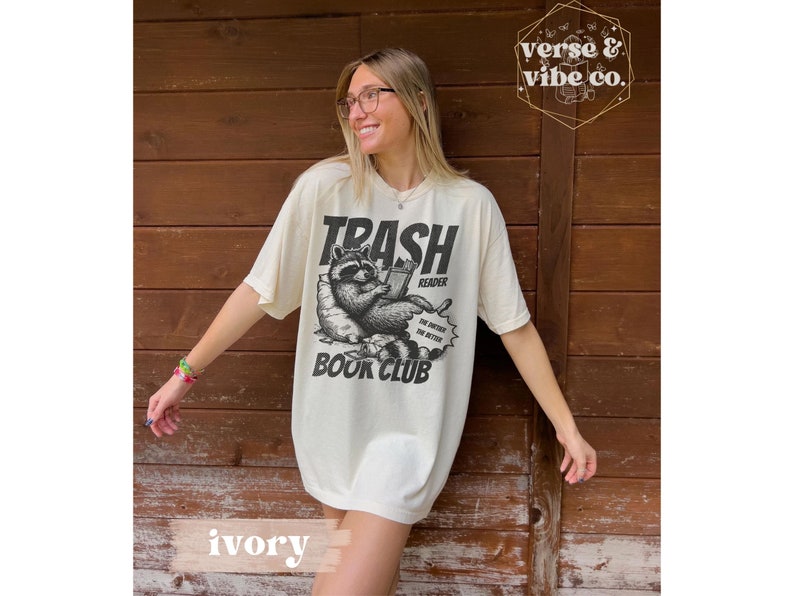 Puede incluir: Una mujer lleva una camiseta de color crema con una ilustraci&oacute;n en blanco y negro de un mapache leyendo un libro. La camiseta dice "Trash Reader Book Club" y tiene un bocadillo que dice "The dirtier the better".