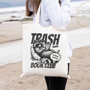 Puede incluir: Una bolsa de tela blanca con una ilustraci&oacute;n en blanco y negro de un mapache leyendo un libro. El texto en la bolsa dice "Trash Reader Book Club" y "The dirtier, the better".