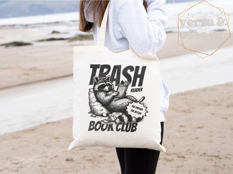 Puede incluir: Una bolsa de tela blanca con una ilustraci&oacute;n en blanco y negro de un mapache leyendo un libro. El texto en la bolsa dice "Trash Reader Book Club" y "The dirtier, the better".