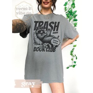 Puede incluir: Camiseta gris con un gr&aacute;fico de un mapache leyendo un libro. El texto en la camiseta dice "Trash Reader Book Club" y "The dirtier the better".