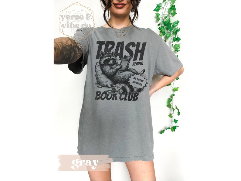 Puede incluir: Camiseta gris con un gr&aacute;fico de un mapache leyendo un libro. El texto en la camiseta dice "Trash Reader Book Club" y "The dirtier the better".