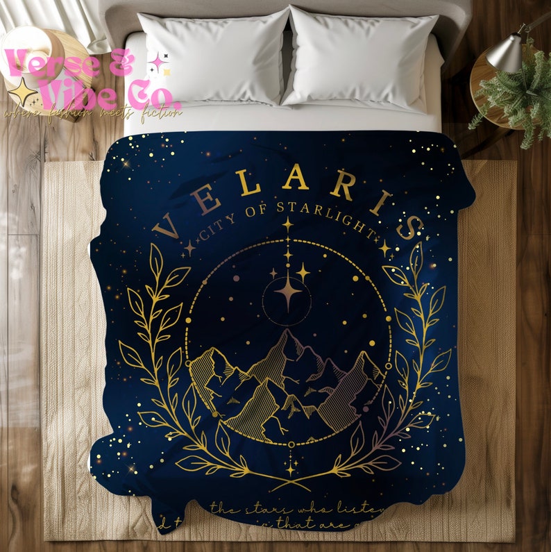 K&ouml;nnte beinhalten: Eine marineblaue Tagesdecke mit einem goldenen Design, das eine Bergkette, Sterne und den Text "VELARIS CITY OF STARLIGHT" zeigt.