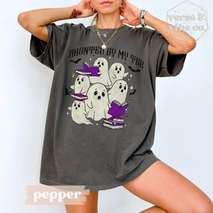 May include: Dark grey t-shirt featuring a graphic of cartoon ghosts with books, and the text "HAUNTED BY MY TBR". The design includes purple books and bats, ideal for book lovers and Halloween.