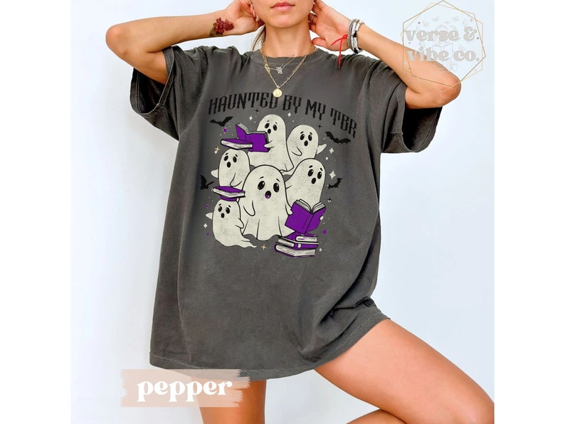 May include: Dark grey t-shirt featuring a graphic of cartoon ghosts with books, and the text "HAUNTED BY MY TBR". The design includes purple books and bats, ideal for book lovers and Halloween.