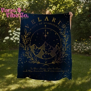 K&ouml;nnte beinhalten: Eine dunkelblaue Wolldecke mit einem goldenen Design, das eine Bergkette, Sterne und den Text "VELARIS - CITY OF STARLIGHT" zeigt. Der Text "To the stars who listen and the dreams that are answered" ist ebenfalls enthalten.