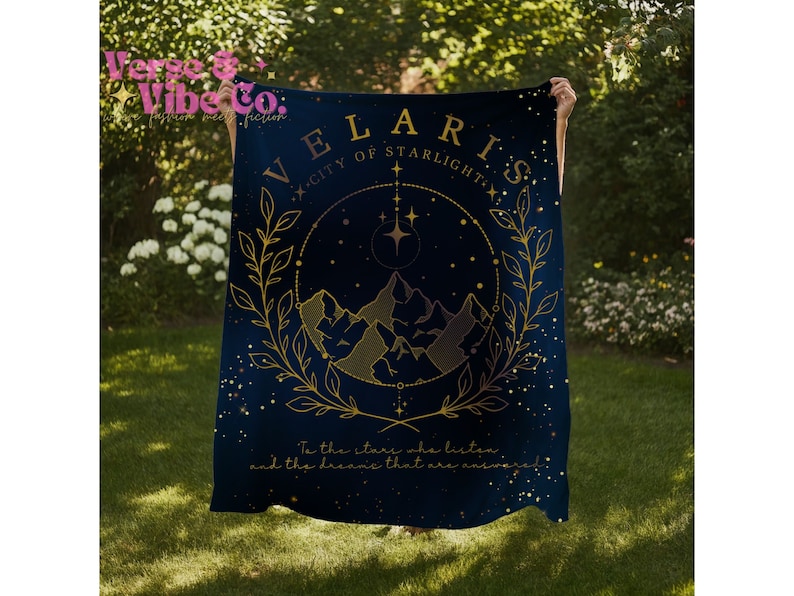 K&ouml;nnte beinhalten: Eine dunkelblaue Wolldecke mit einem goldenen Design, das eine Bergkette, Sterne und den Text "VELARIS - CITY OF STARLIGHT" zeigt. Der Text "To the stars who listen and the dreams that are answered" ist ebenfalls enthalten.