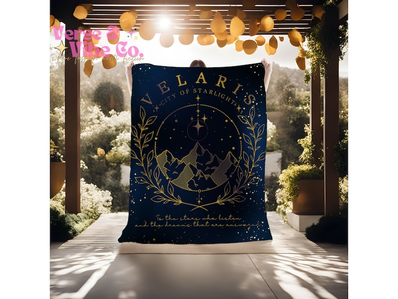 K&ouml;nnte beinhalten: Eine marineblaue Kuscheldecke mit einem goldenen Design, das eine Bergkette, Sterne und den Text "VELARIS - CITY OF STARLIGHT" zeigt. Der Text "To the stars who listen and the dreams that are whispered" befindet sich am unteren Rand der Decke.