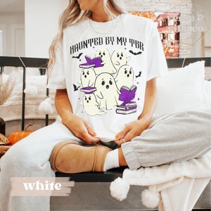 May include: White t-shirt featuring the text "HAUNTED BY MY TBR" and a graphic of cartoon ghosts with books. The design includes purple book accents and bat details, ideal for Halloween or book enthusiasts.