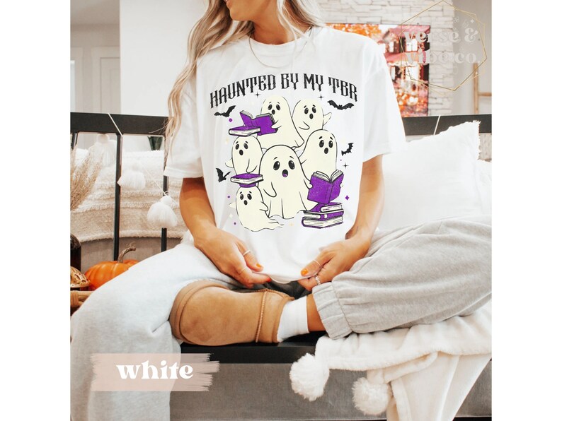 May include: White t-shirt featuring the text "HAUNTED BY MY TBR" and a graphic of cartoon ghosts with books. The design includes purple book accents and bat details, ideal for Halloween or book enthusiasts.