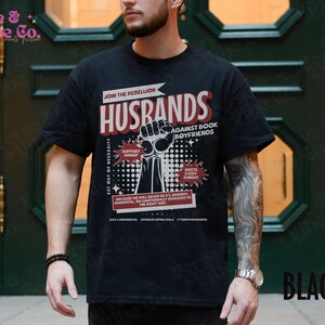 以下が含まれることがあります： 「Join the Rebellion: Husbands Against Book Boyfriends」というテキストと、拳を突き上げたグラフィックがプリントされた黒いTシャツ。グラフィックには、「Support Group」、「Meets Every Sunday」、「Est Out of Necessity」というテキストも含まれています。