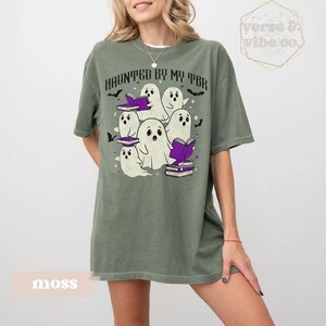 May include: Moss green t-shirt featuring the text "HAUNTED BY MY TBR" and a graphic of cartoon ghosts. The ghosts are white with black eyes and are wearing purple book hats. The shirt has a relaxed fit.