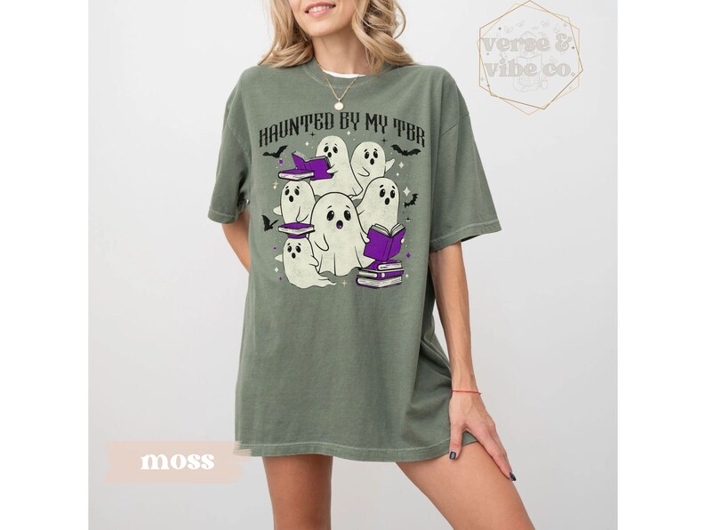 May include: Moss green t-shirt featuring the text "HAUNTED BY MY TBR" and a graphic of cartoon ghosts. The ghosts are white with black eyes and are wearing purple book hats. The shirt has a relaxed fit.