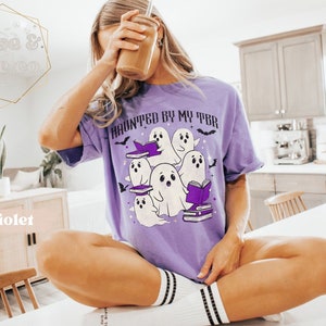 May include: A lilac t-shirt featuring the phrase "HAUNTED BY MY TBR" and a design of cartoon ghosts, books, and bats. The ghosts are white with black eyes and are holding books. The shirt is a relaxed fit.