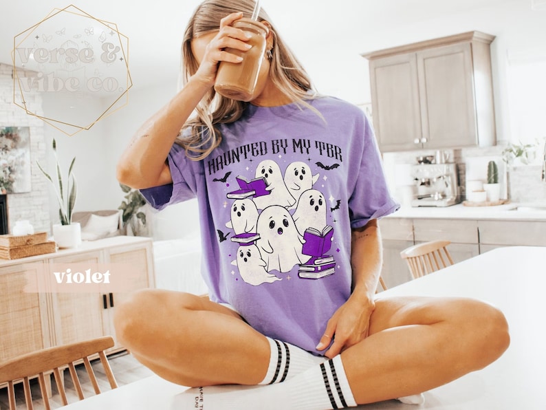 May include: A lilac t-shirt featuring the phrase "HAUNTED BY MY TBR" and a design of cartoon ghosts, books, and bats. The ghosts are white with black eyes and are holding books. The shirt is a relaxed fit.